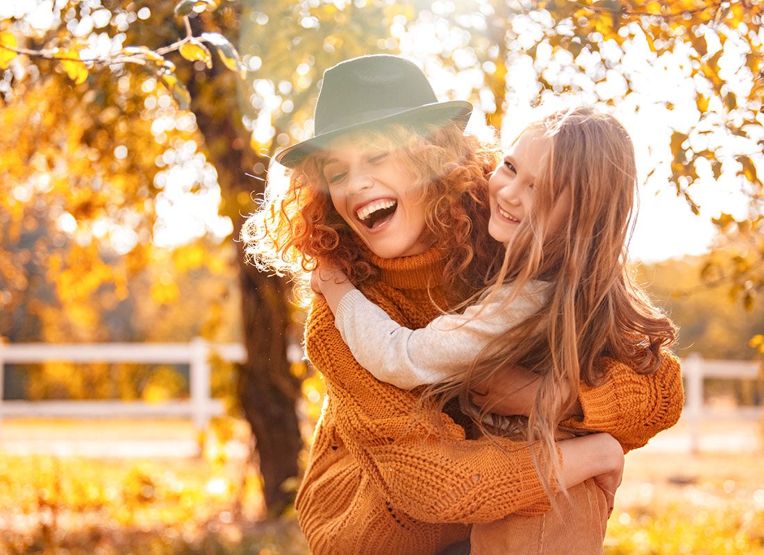 About Our Agency - Closeup Portrait of a Joyful Young Mother Hugging Her Daughter While They Enjoy a Beautiful Sunny Day in the Countryside During the Fall